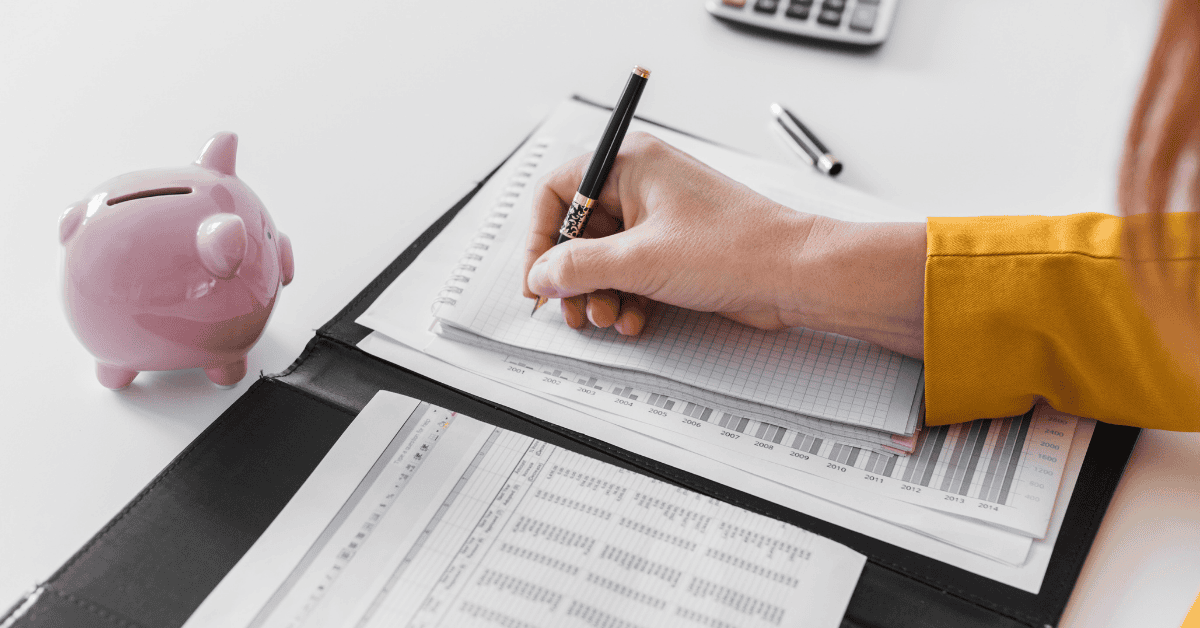 Mão escrevendo em planilha financeira ao lado de um cofrinho rosa, organizando orçamento para solicitar um empréstimo.