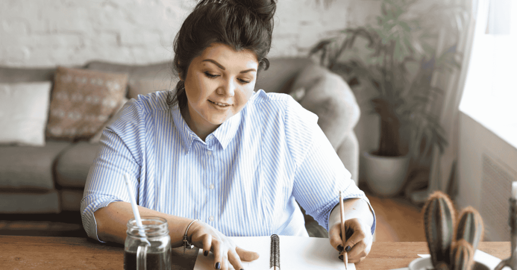 Mulher sentada à mesa escrevendo em um caderno, com uma bebida ao lado, aparentando planejar finanças para solicitar um empréstimo.