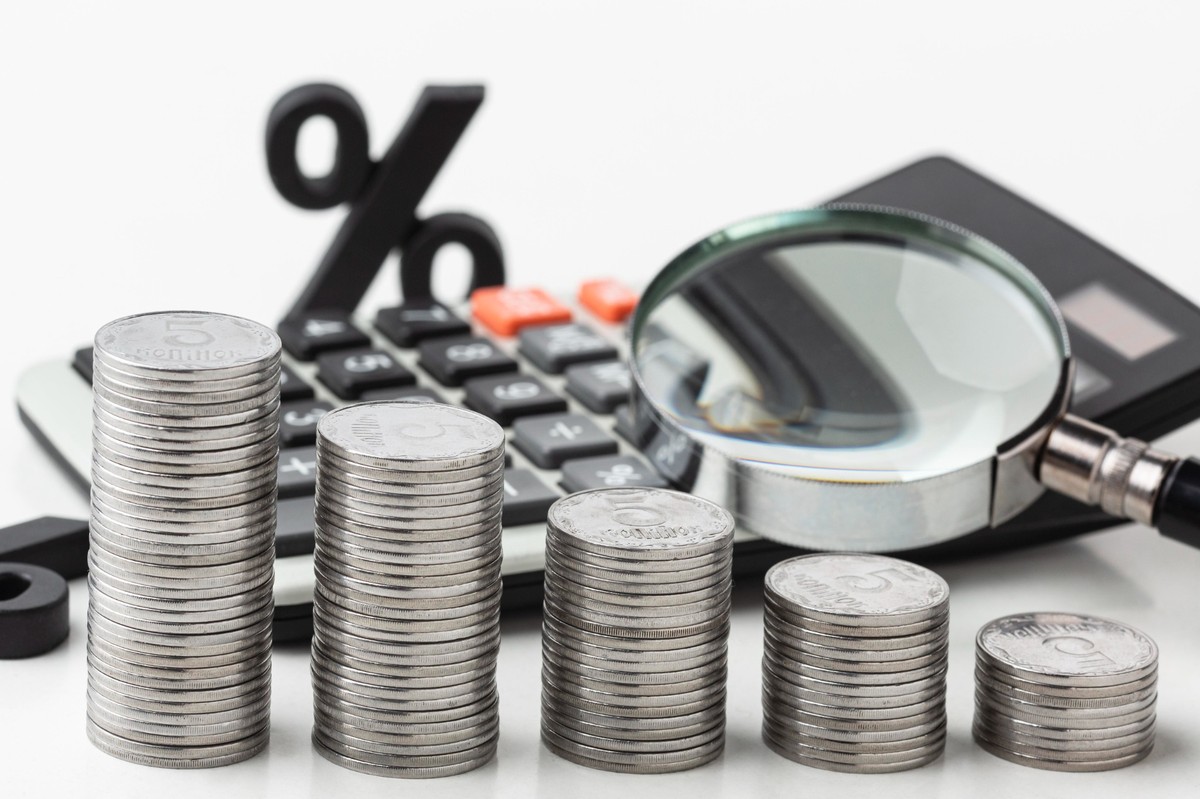 “Fotografia de pilhas de moedas prateadas organizadas em ordem crescente, posicionadas à frente de uma calculadora e de uma lupa, com um símbolo percentual desfocado ao fundo. A composição remete à análise detalhada de custos e juros, destacando visualmente o impacto da inflação, que faz o valor do dinheiro diminuir ao longo do tempo. A lupa reforça a necessidade de examinar preços, reajustes e variações econômicas com atenção.
