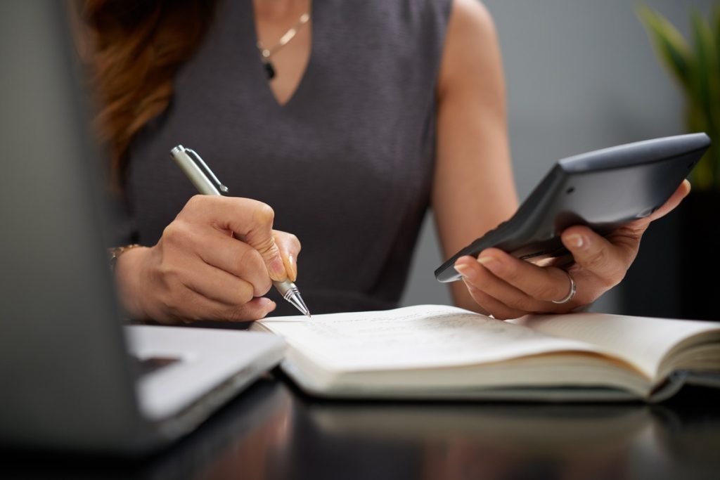 Mulher fazendo anotações em um caderno enquanto segura uma calculadora, com um laptop aberto à frente. A imagem mostra atenção aos números e registros, simbolizando o processo de acompanhar despesas fixas e variáveis. A composição reforça a importância do controle financeiro, destacando o hábito de revisar valores, categorizar gastos e monitorar o orçamento para manter equilíbrio nas finanças.